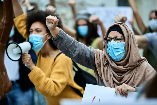 Muslim woman with raised fist wearing protective face mask while taking a part in public demonstrations. 