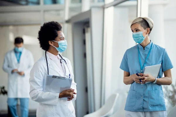 Black female doctor and nurse with face masks communicating while standing in a hallway at medical clinic.
