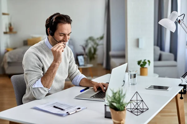 Happy entrepreneur wearing headset and talking to someone while working on laptop at home