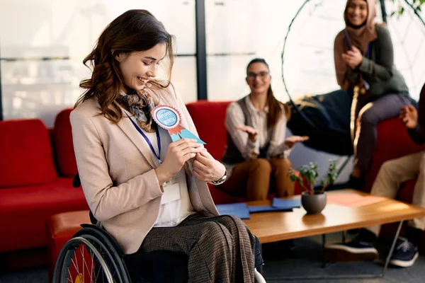 Happy disabled businesswoman receiving employee of the month badge while working at corporate office. Group of colleagues are applauding her in the background.