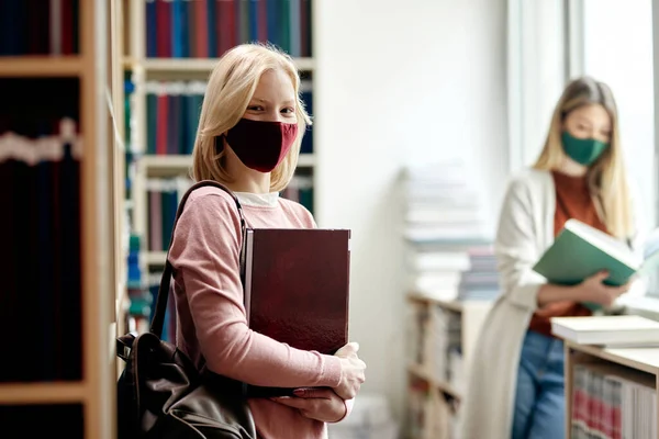 Happy female student holding books and wearing face mask in a library while looking at camera.