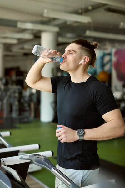 Thirsty athletic man refreshing himself with water after running on treadmill at health club.