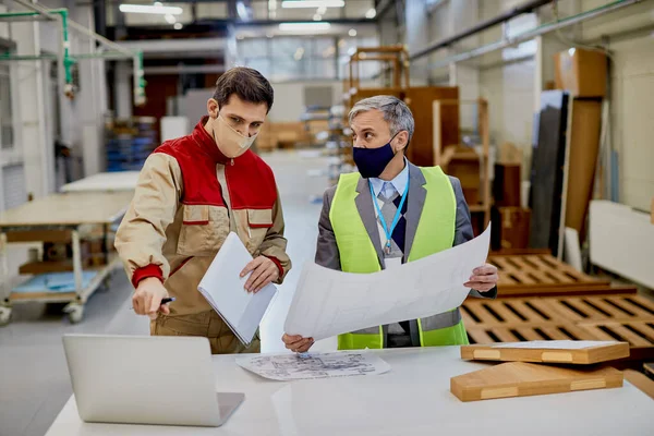 Woodworking engineer and male worker using laptop while analyzing blueprints in a factory during coronavirus pandemic.