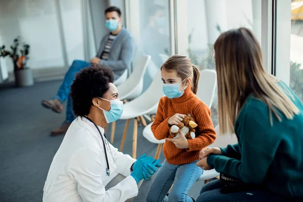 Little girl and her mother communicating with black female doctor in a waiting room at medical clinic. They are wearing face masks due to COVID-19 pandemic.