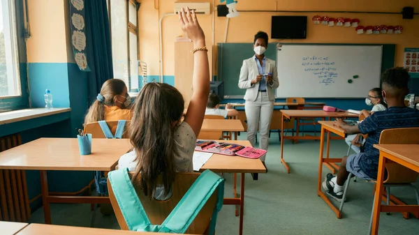 Back view of elementary student raising her hand to answer teacher#s question during a class at school.