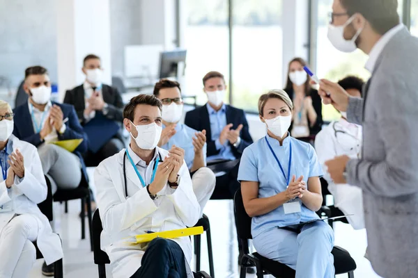 Large group of doctors and business people with protective face masks applauding while attending an educational event at conference hall. Focus is on male doctor.