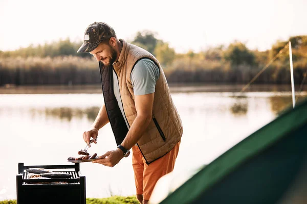 Happy mid adult man grilling meat on barbecue during his camping day in nature.
