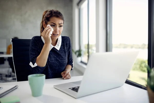 Sad businesswoman using computer and crying while talking to someone via video call from her office.