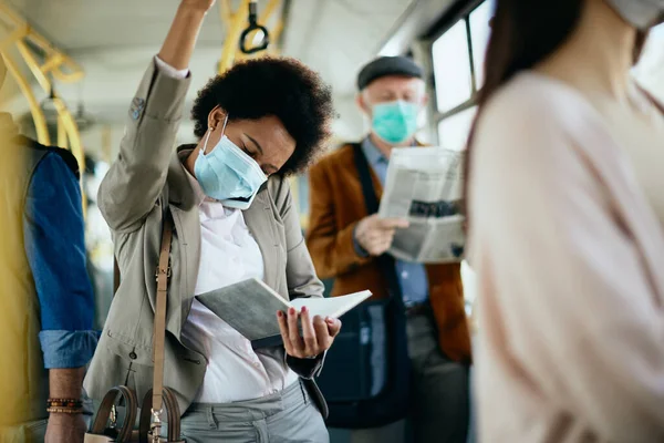 Black businesswoman with face mask reading notes while talking on the phone and commuting by public transport.