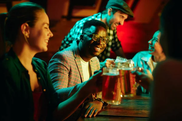 Young African American man and his friends toasting with beer in a pub at night.