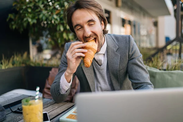 Young happy businessman using laptop and eating croissant on a break in outdoor cafe.