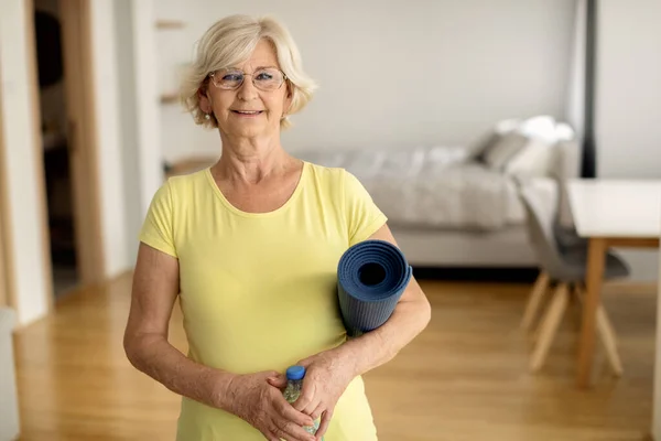 Happy mature woman getting ready for home exercising and holding exercise man and bottle of water while looking at camera.