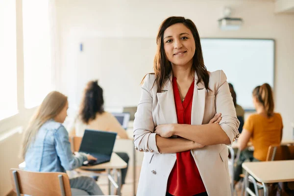 Happy female professor standing with arms crossed during computer class and looking at camera. Her students are e-learning in the background.