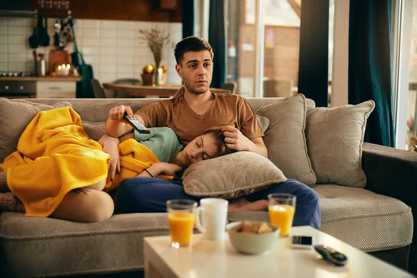 Young man watching TV and changing channels while girlfriend is napping on his lap at home.