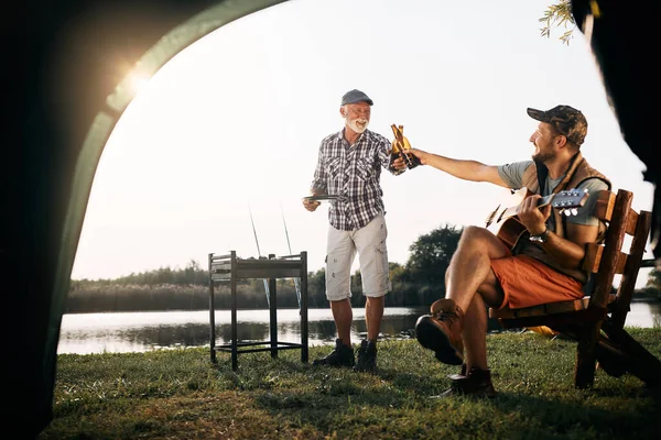 Happy man playing acoustic guitar and toasting with his father who is grilling meat during their camping day.