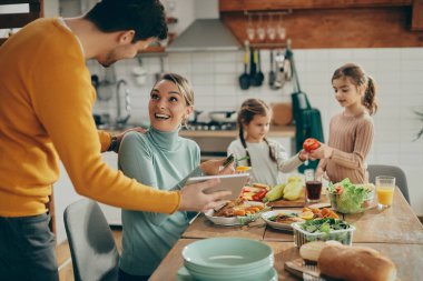 Happy couple having fun while using touchpad at dining table while their daughters are in the background. Focus s on woman. 