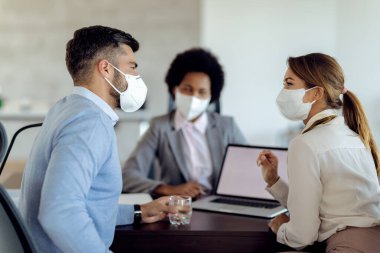 Happy couple with protective face masks communicating while having a meeting with real estate agent in the office. 