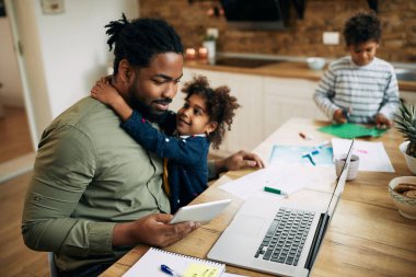 Happy African American little girl embracing her father while he's working on laptop and digital tablet at home. 