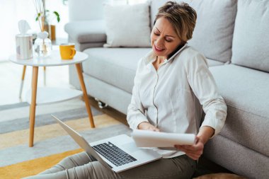 Happy businesswoman talking on the phone while using laptop and reading reports at home. 