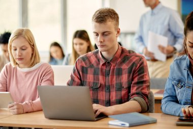 Young student using laptop while attending a class with his friends at college classroom.