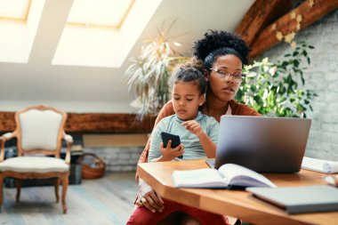 African American stay at home mother working on laptop while daughter is sitting on her lap and using smart phone.