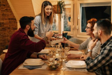 Young happy woman serving dinner to her friends at dining table. 