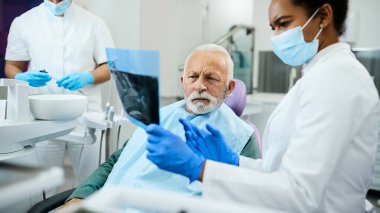 African American dentist and her senior patient examining orthopantomogram at dental clinic.