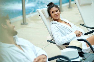 Young couple in bathrobes talking while relaxing by the pool at the spa. Focus is on woman.