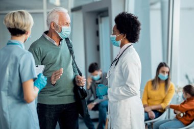 Senior man and black female doctor with protective face masks talking in a hallway at medical clinic. 