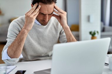 Exhausted entrepreneur holding his head in pain after working on a computer at home.