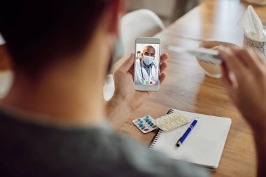 Close-up of man measuring his body temperature and having video call with his doctor over mobile phone. Focus is on doctor on touchscreen. 
