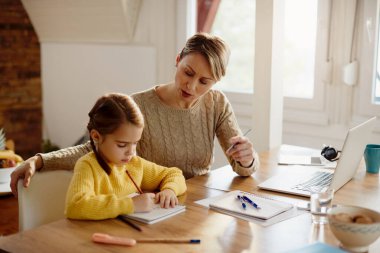 Working mother talking to her small daughter who is drawing in notebook while homeschooling. 