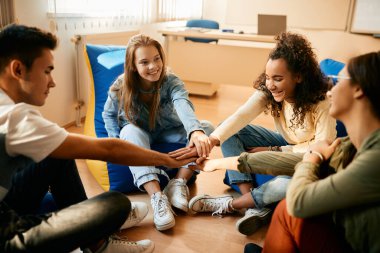 Multiracial group of teenagers uniting hands while celebrating their friendship in the classroom.