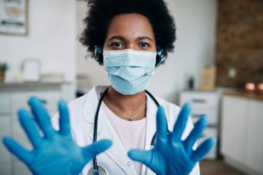 Black female doctor wearing protective face mask and gloves while having conference call and looking at camera.