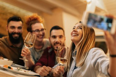 Cheerful woman having fun while drinking wine and taking selfie with her friend in dining room. 