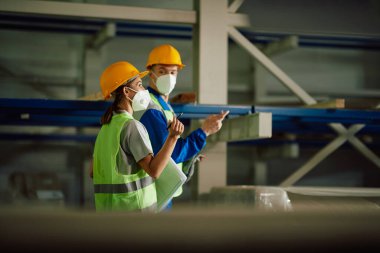 Two warehouse workers communicating while walking through storage room and wearing protective face masks. 