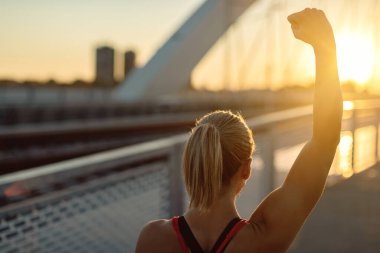 Back view of sportswoman with raised arm celebrating successful training at sunset. 