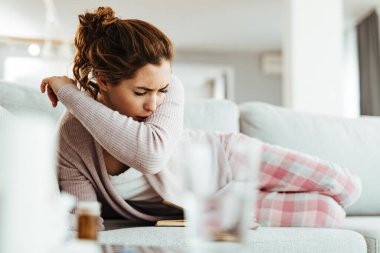 Young woman coughing into elbow while lying down on sofa in the living room.