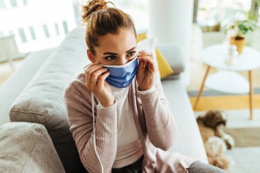 Young woman using protective face mask while sitting on the sofa at home. 
