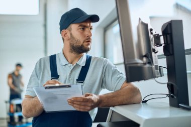 Otomobil tamircisinde çalışırken masaüstü bilgisayarı kullanan ve notlar yazan araba tamircisi.