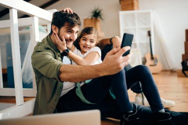 Playful single father and his daughter having fun while taking selfie at their new apartment. 