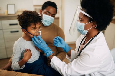 African American pediatrician using nebulizer during inhaling therapy of a small boy due to coronavirus pandemic. 