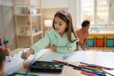 Creative small girl using water colors while painting at kindergarten. 
