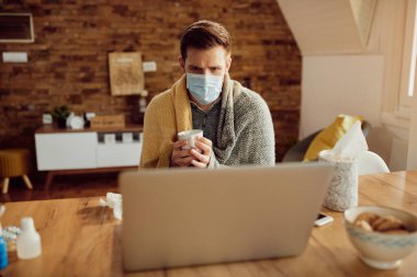 Sick man wrapped in a blanket having cup of tea while using computer and wearing face mask at home.