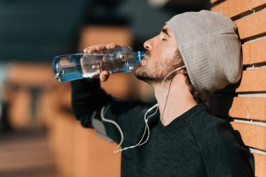 Profile view of exhausted athletic man drinking fresh water from a bottle while leaning on a wall outdoors. 