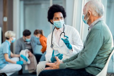 Black female doctor and senior man wearing protective face masks while communicating in a waiting room at hospital. 