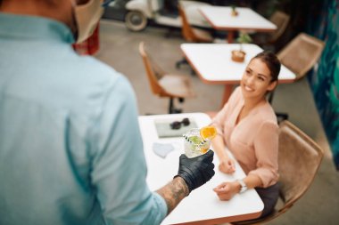 Close-up of waiter wearing protective glove while serving drink to female guest at reopened cafe.