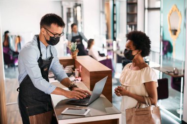 Male hairstylist using laptop while talking to African American woman at reception desk. They are wearing face masks due to COVDI-19 pandemic. 