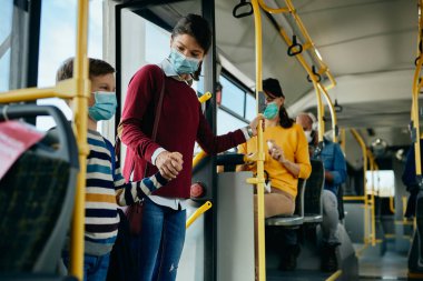 Happy mother and son holding hands and wearing face masks while entering in a bus.