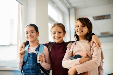 Low angle view of little girls standing embraced in the classroom at elementary school and looking at camera. 